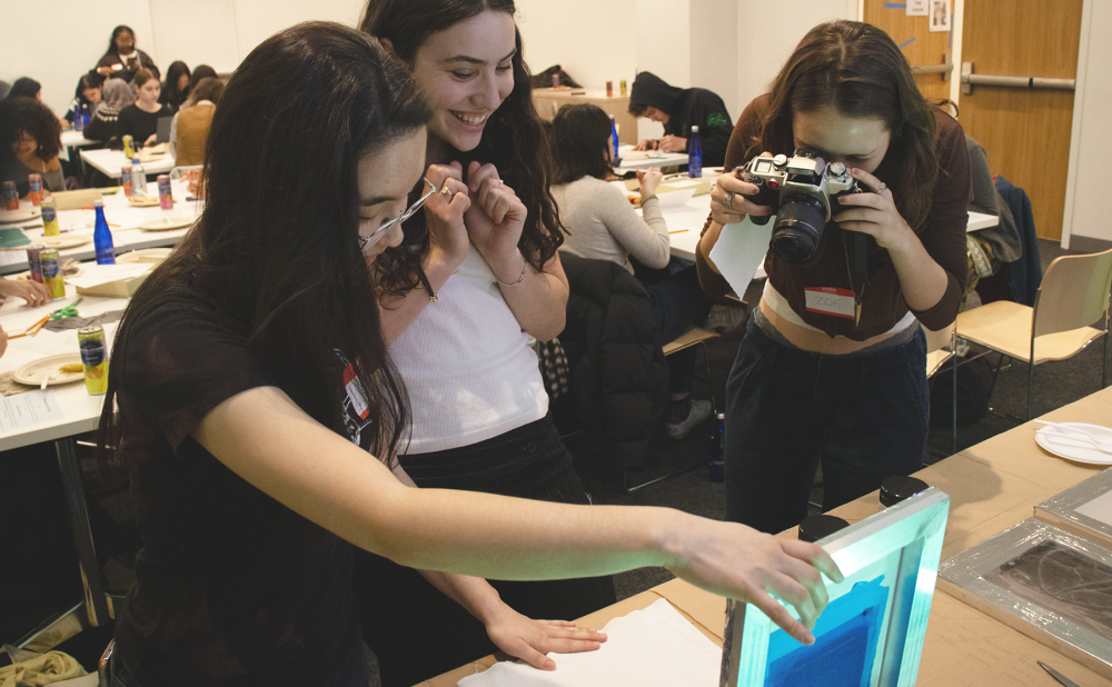 Three women engaged in a screen printing activity, with one taking a photograph of the process.