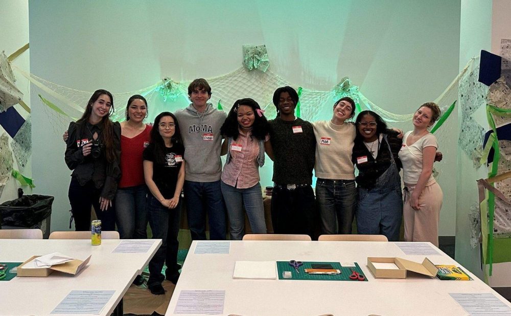 A group of ten people posing in front of artistic wall installations in a bright room with crafting tables in the foreground. Alt-text generated by AI.