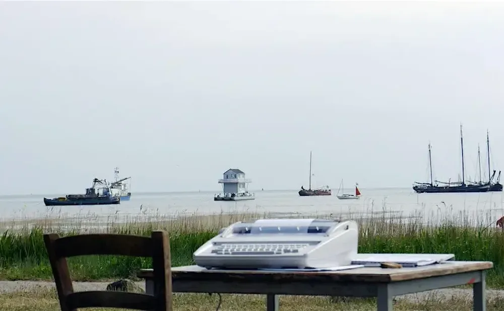 A modern typewriter sits on a table outside in front of grass, a lake with boats, beneath a white sky.