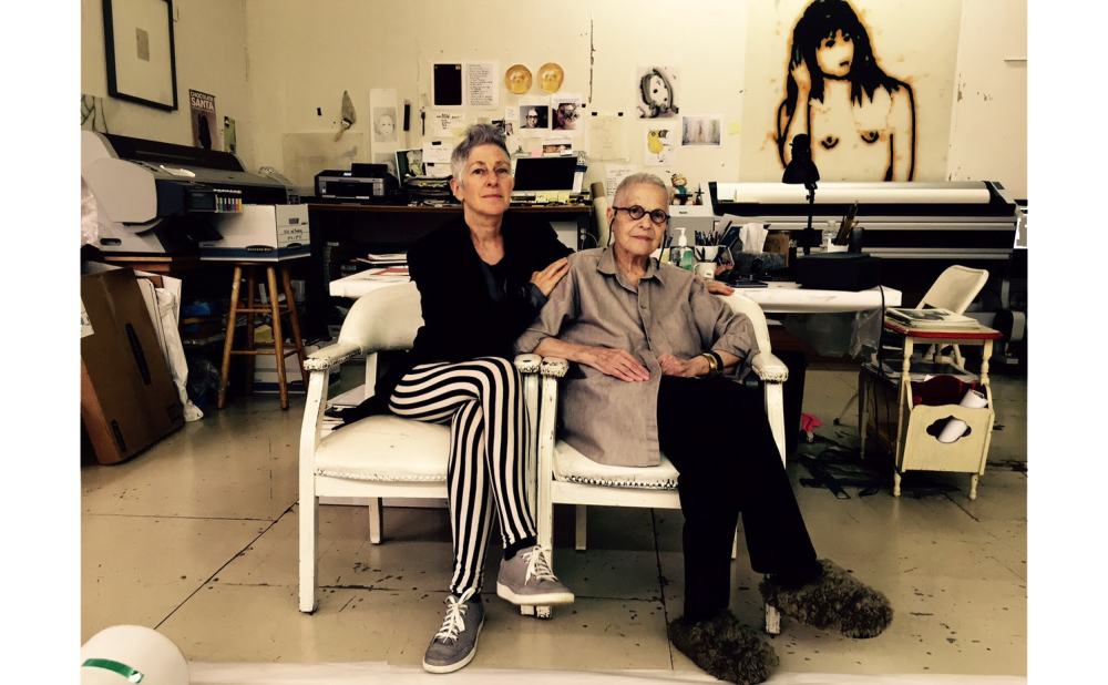 Two women sitting next to each other on white chairs in an art studio with various artworks and a large printer behind them.