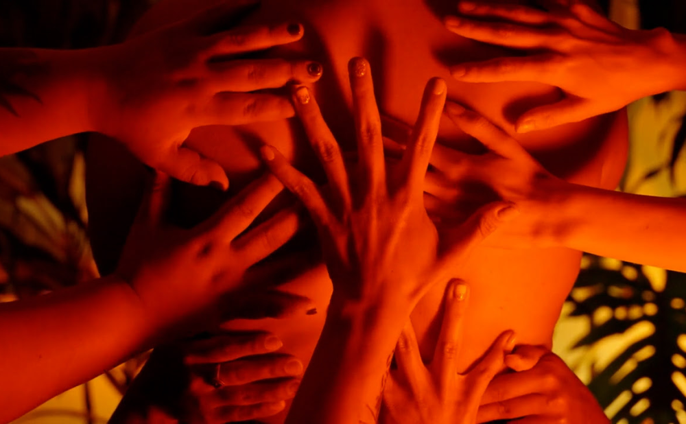 Film still showing an individual with their back facing the camera, with seven hands spread open on their back. The individuals are bathed in yellow and deep orange light.