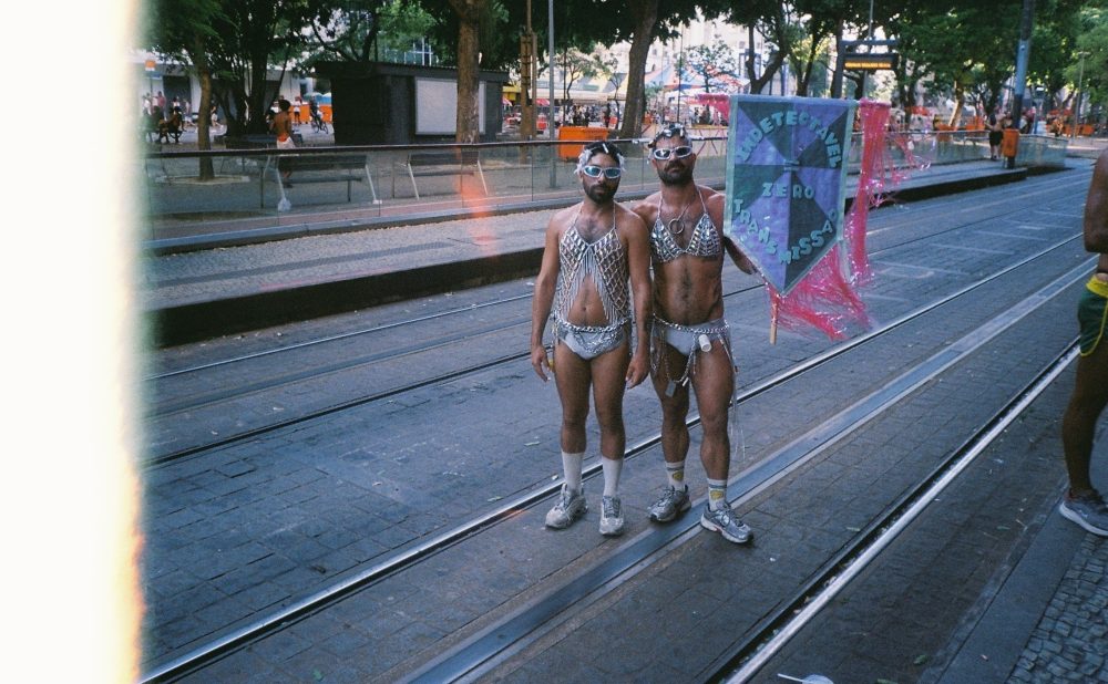 Two individuals in silver chain outfits standing on tram tracks, one holding a banner.