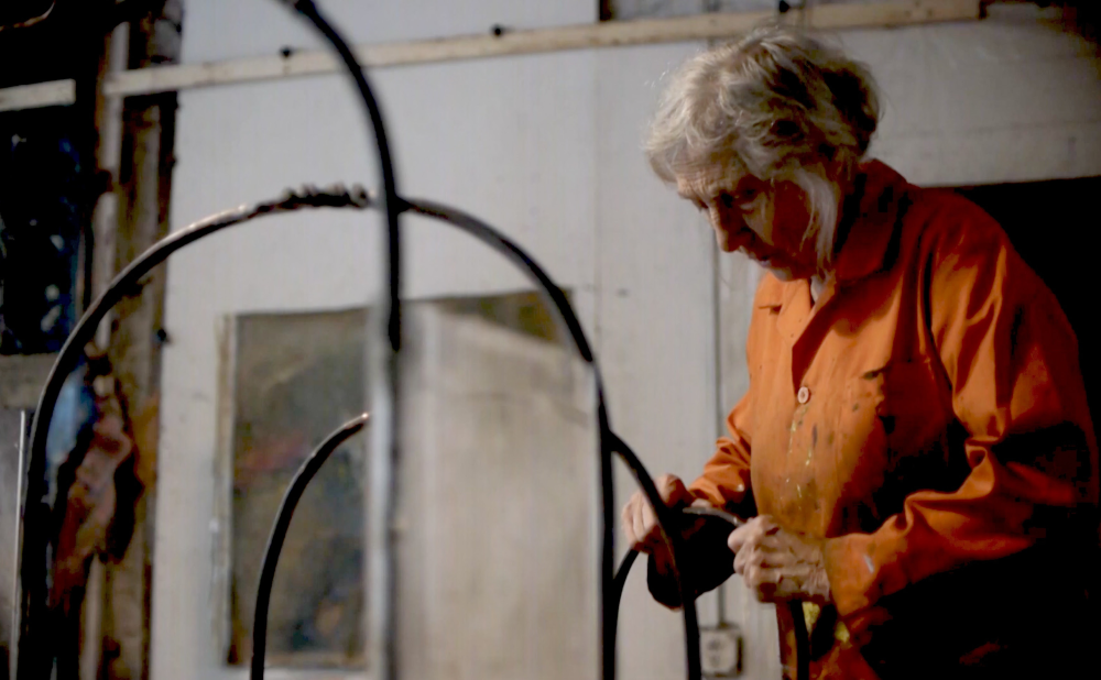 An older woman with white hair in an orange jumpsuit working on a metal sculpture inside an art studio.