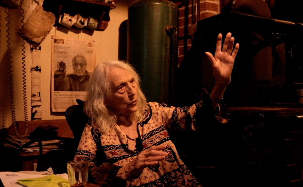 An older woman with medium length white hair in a dimly lit room, raising an arm and gesturing.