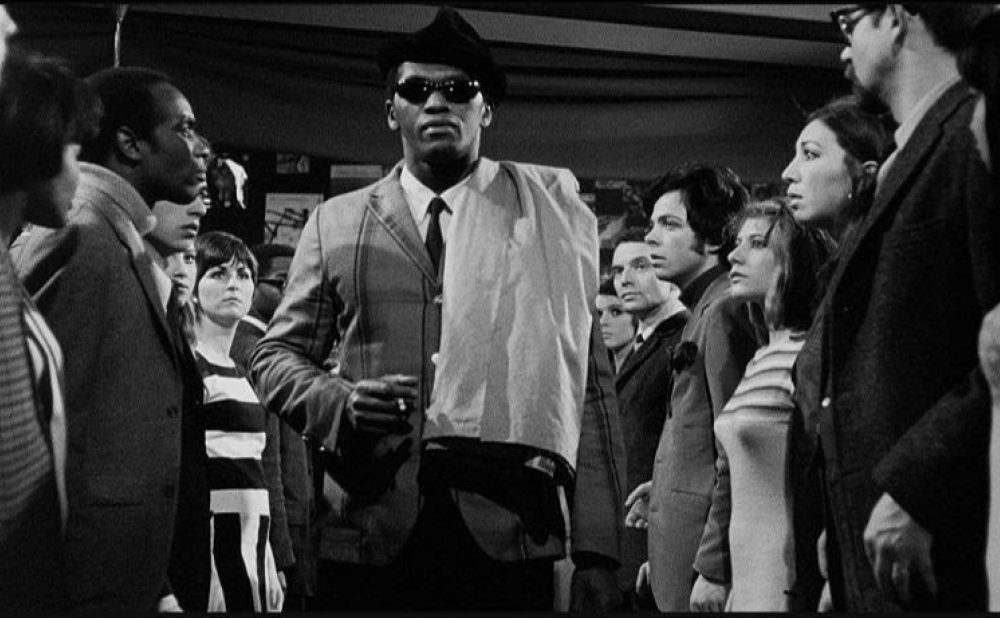 Black-and-white film screencap showing a black man in a suit with hat and dark glasses striding toward the camera, cigarette in hand; he is flanked by rows of people staring at him in a serious manner.