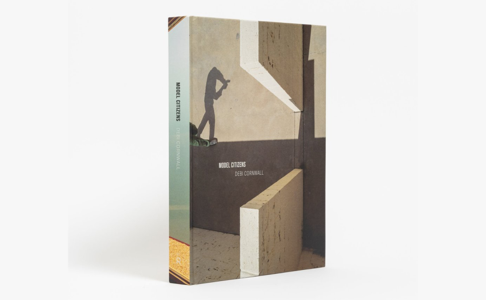 Book standing upright on a white background, with a cover showing a person's shadow in an industrial looking setting, with title Model Citizens and author Debi Cornwall listed on the cover and spine.
