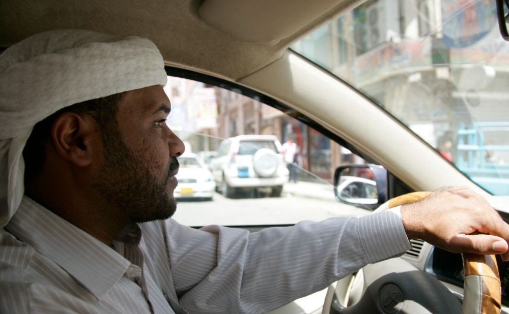 Man driving a car with street scene visible outside the window.