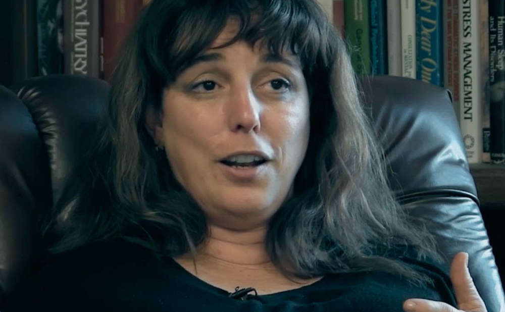 Close-up view of a Cuban woman with long brown hair and bangs sitting in a leather chair, talking to the camera or someone behind it; her gesturing hand is barely visible in the frame.