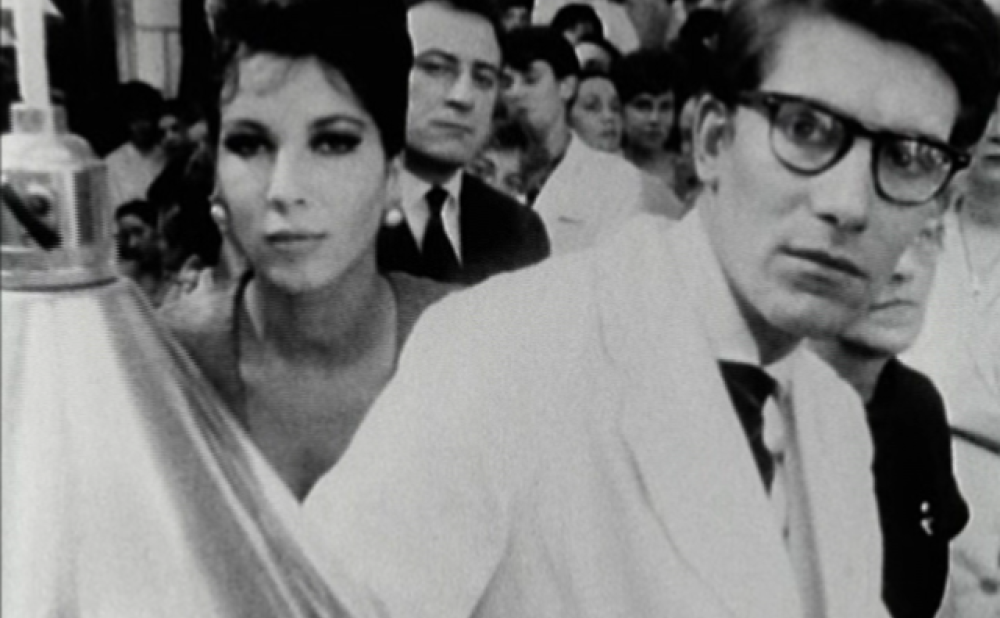 Black-and-white film screencap with a close-up of a bespectacled man in a white suit next to a young woman with a bouffant hairstyle and earings; a crowd of people is visible behind them.