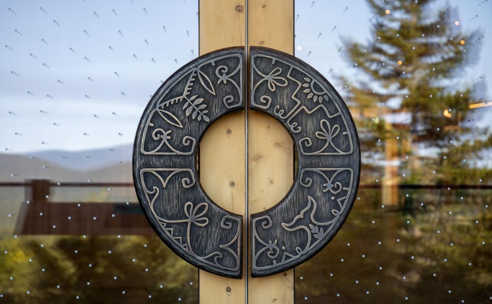 A circular metal door handle with intricate floral carvings on a wooden-framed glass door. The glass reflects a landscape of trees and mountains at the Tekαkαpimək Contact Center.
