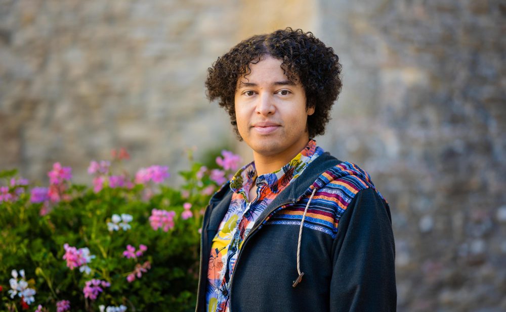 Photograph of Indigenous artist with curly hair and colorful clothing with a floral bush in the background.