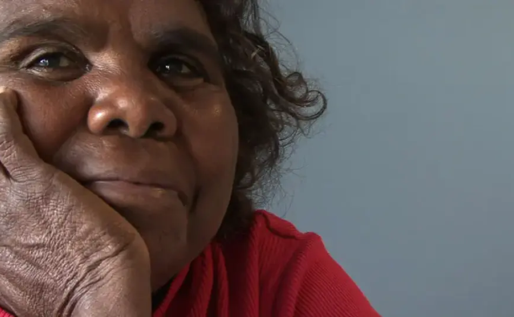 Close-up photograph of a middle-aged Aboriginal woman with her chin resting on her hand, wearing a red shirt.