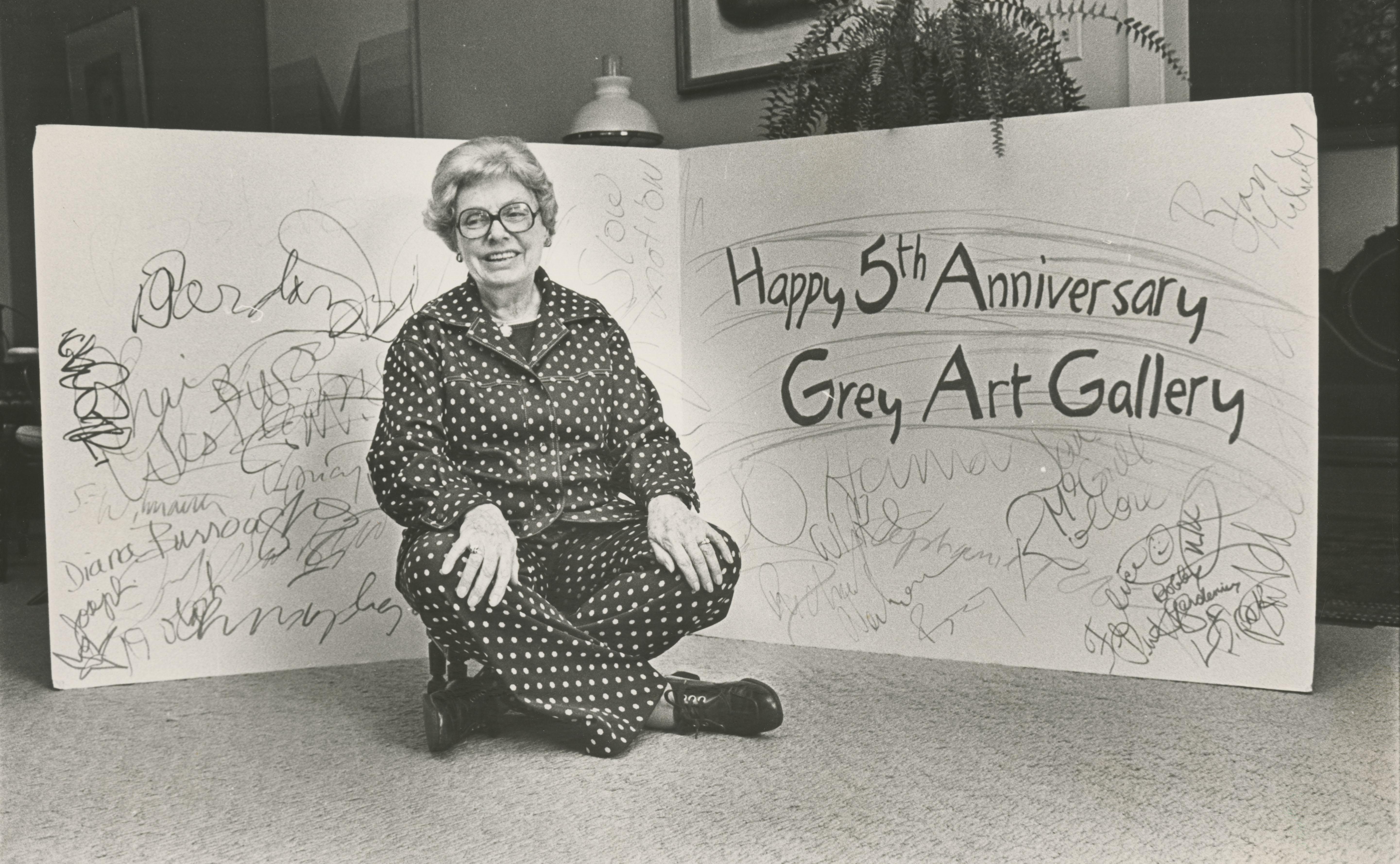An elderly woman with short, wavy gray hair and large, dark-rimmed glasses sitting in front of a signed card marking the 5th anniversary of the Grey Art Gallery, dressed in a polka-dotted outfit.