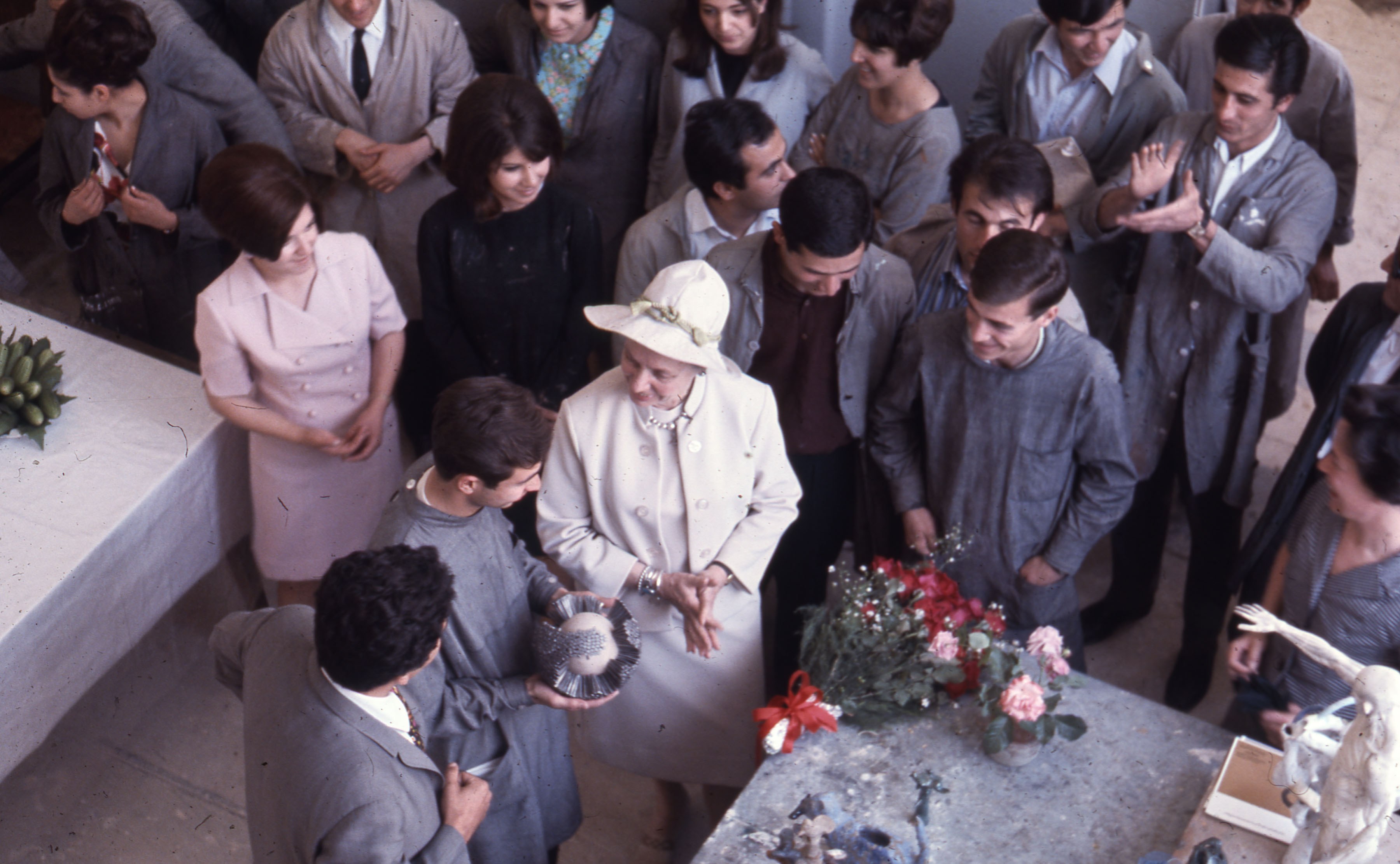 Birds-eye-view photograph of people in a workshop-like environment, with one central woman in a hat observing a sculpture held by a man.