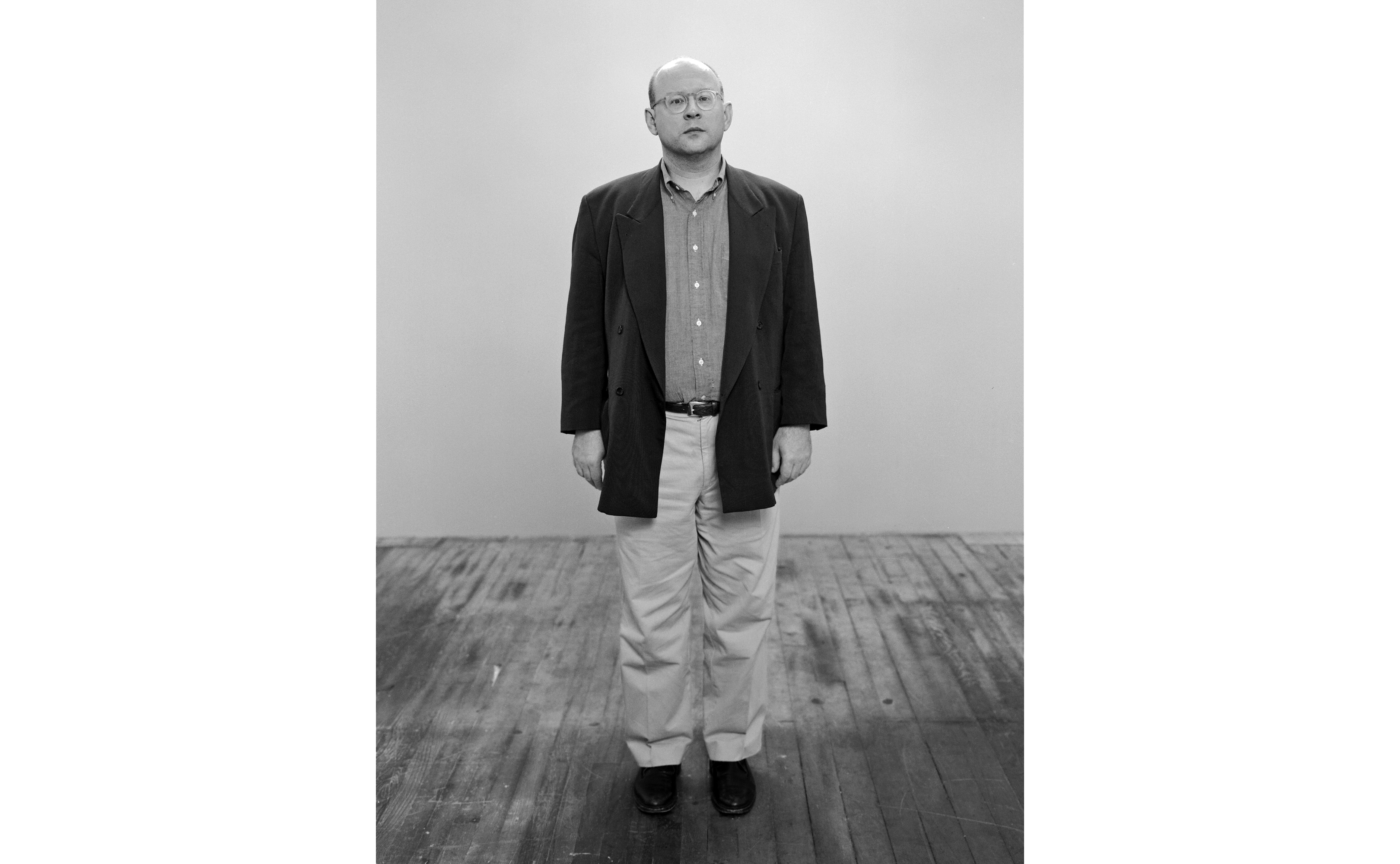 Person standing on a wooden floor, wearing a dark blazer and light pants, against a plain background. Alt-text generated by AI.