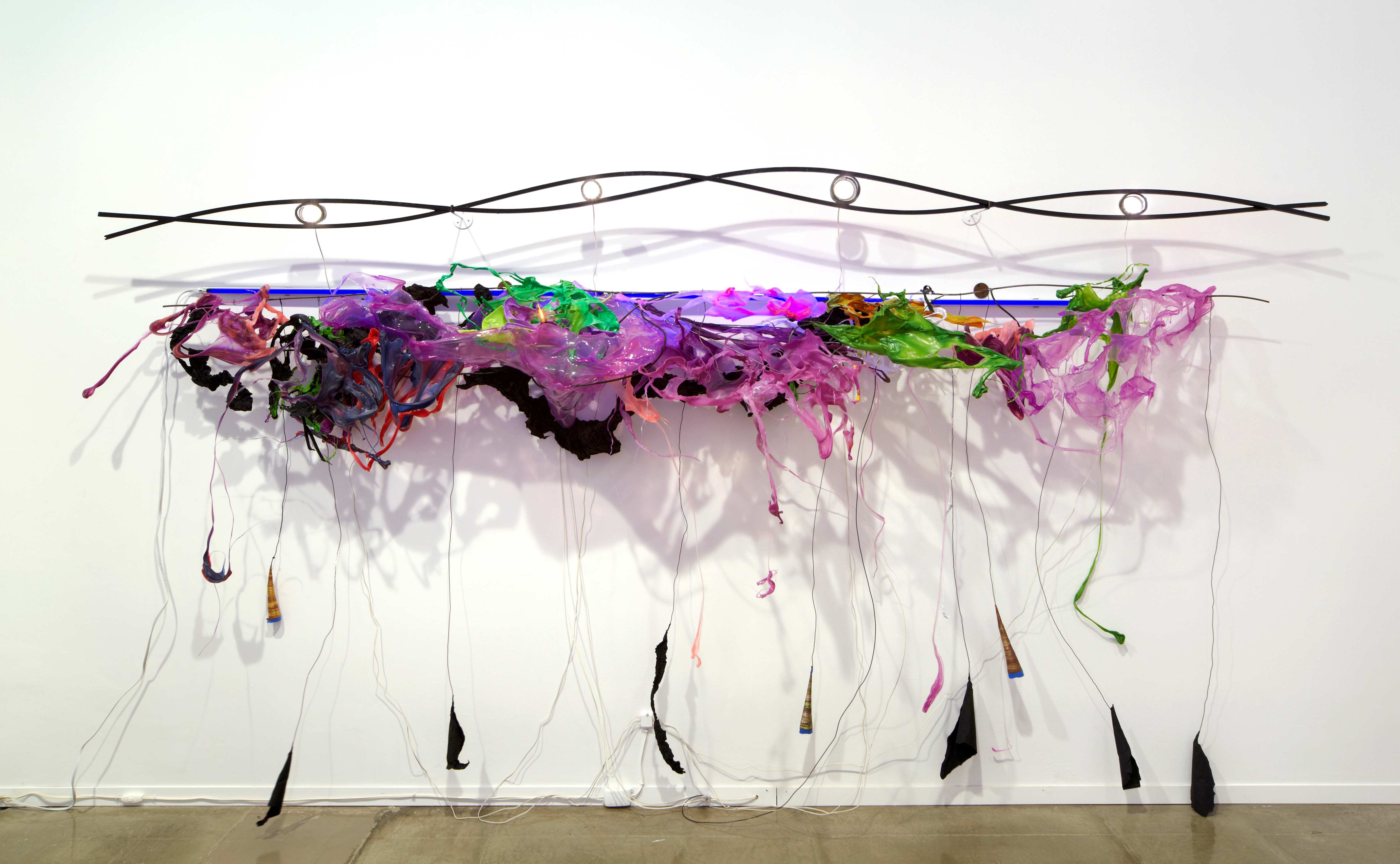 A sculpture comprising of a steel rod twisted in the shape of a DNA strand, hanging above a jubilant array of melted green, pink, and purple plastics and florescent lights. Tendrils of plastics reach down to the gallery floor.