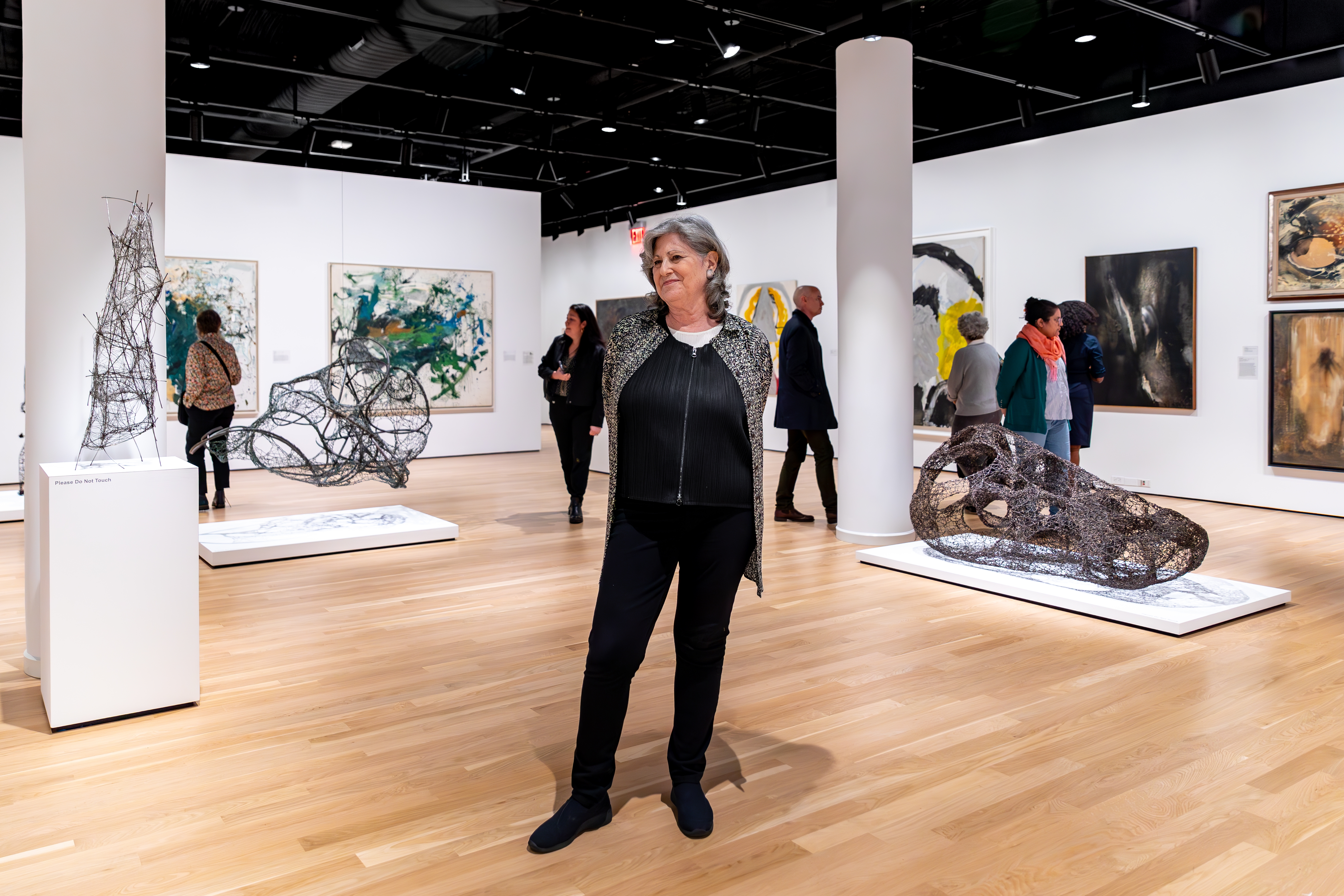 Woman with shoulder length salt and pepper hair standing in the center of an art gallery filled with sculptures and paintings. She stands with her hands behind her back, smiling away from the camera.