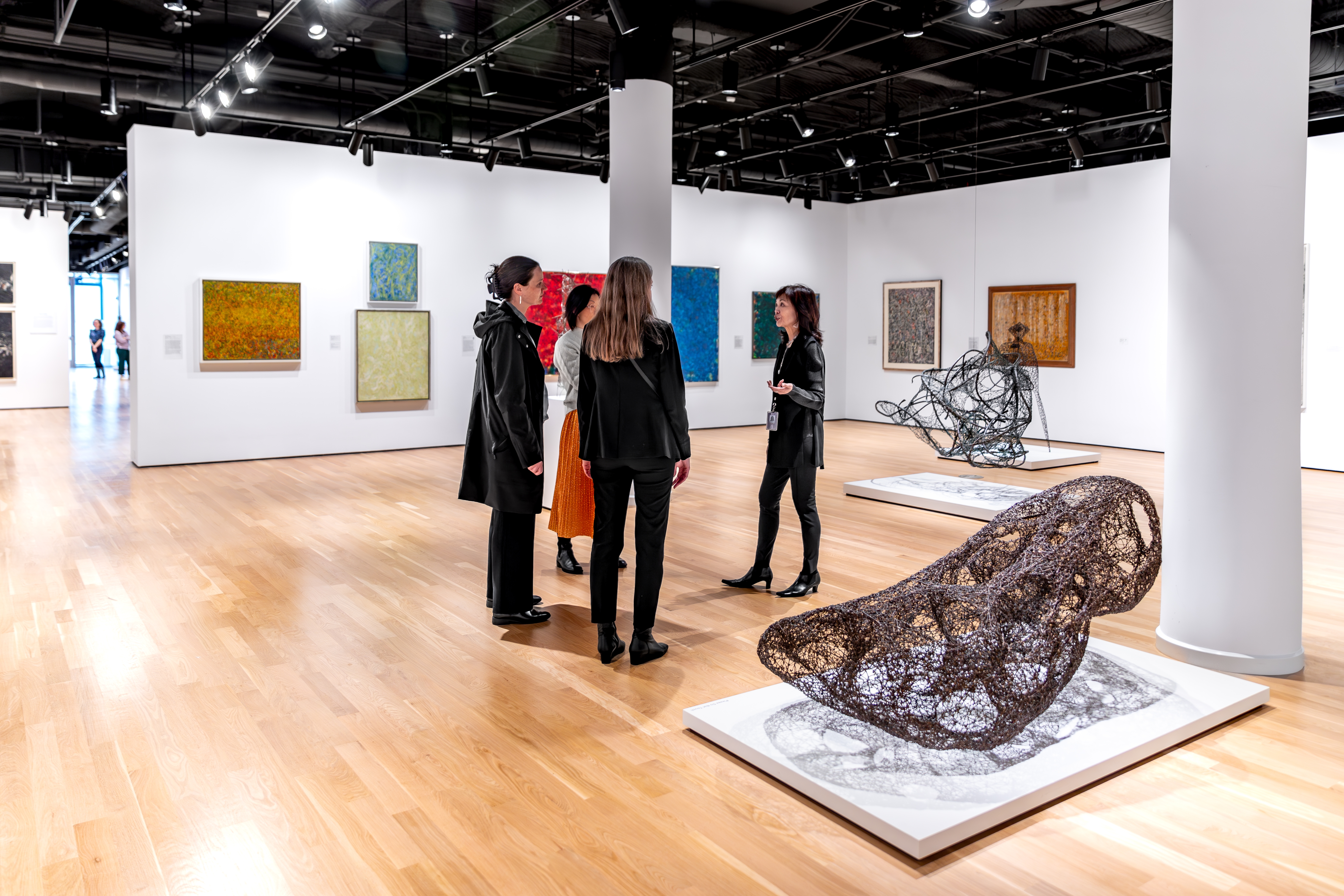 Four women standing and talking in the middle of an art museum; paintings can be seen on the walls in the background along with two metal sculptures, one of which is in the image foreground