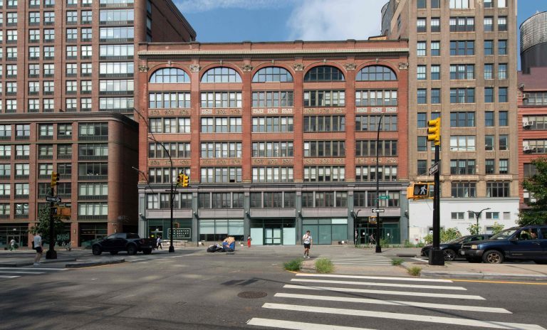 A red-brick building with arched windows centered in a city street scene, flanked by taller buildings.