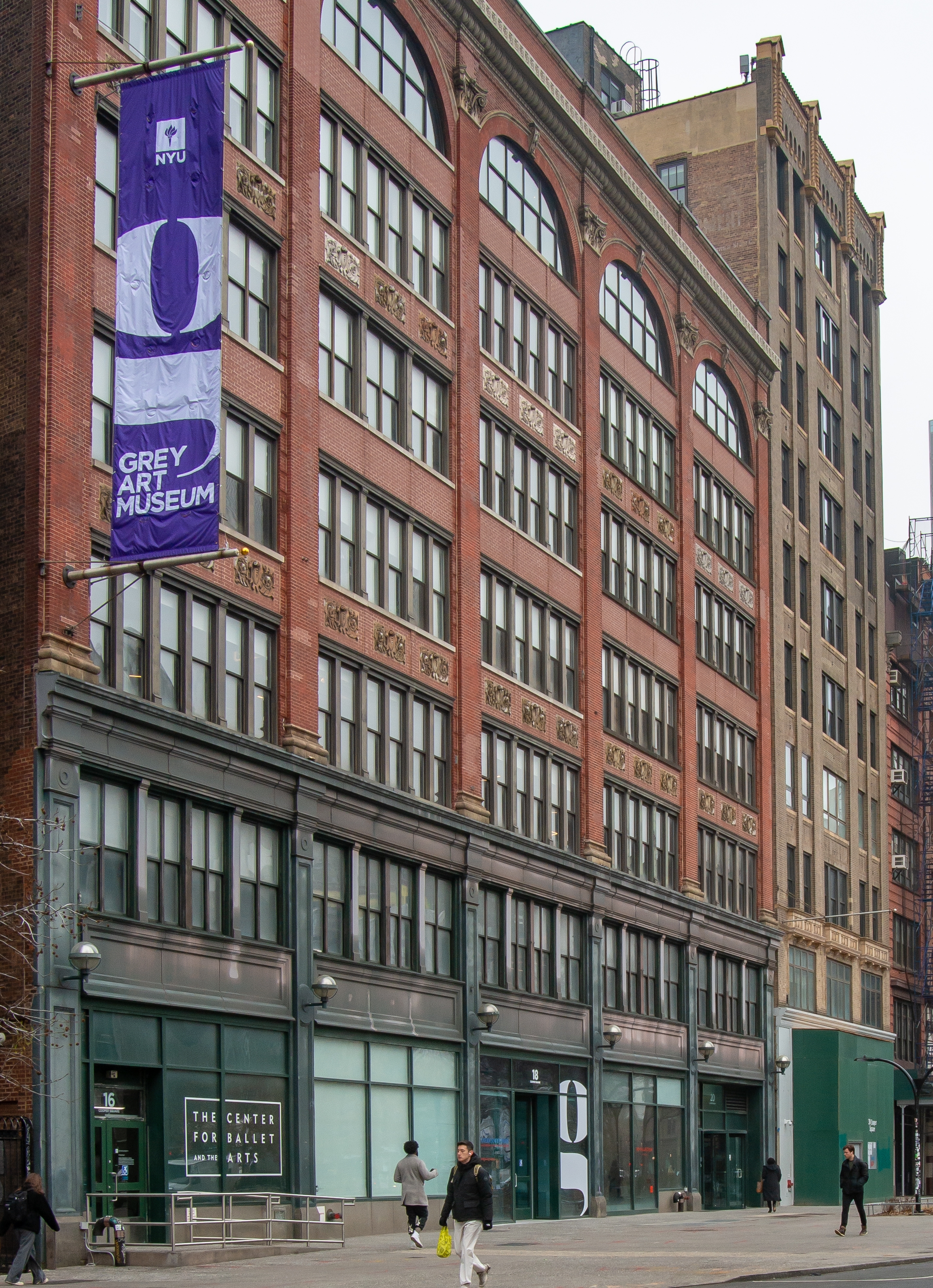 A red brick building with arched windows, decorative stonework, and a vertical violet and white banner displaying NYU and Grey Art Museum.