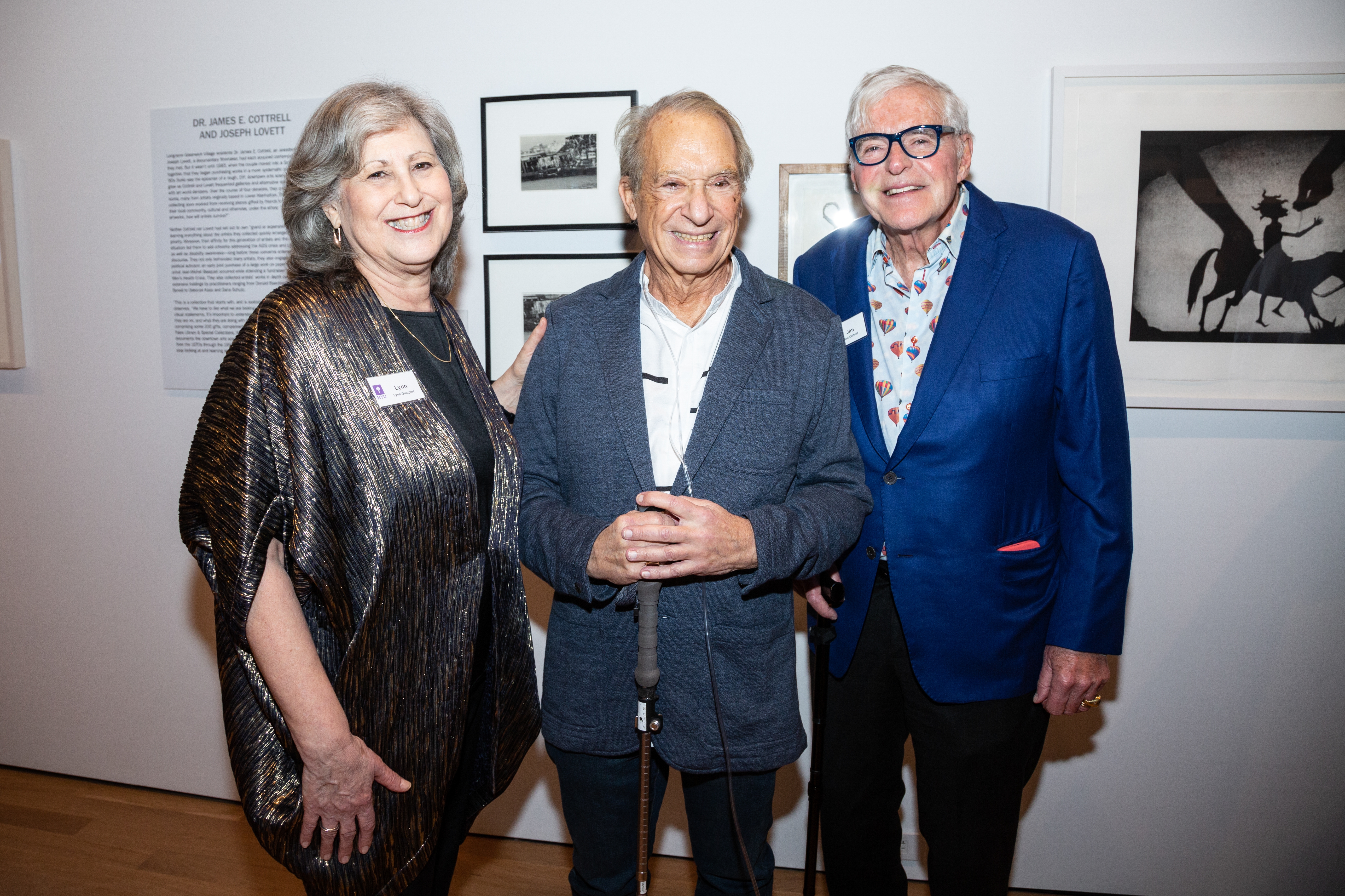 Three people, one older woman and two older men, in a gallery setting stand in front of framed art pieces.